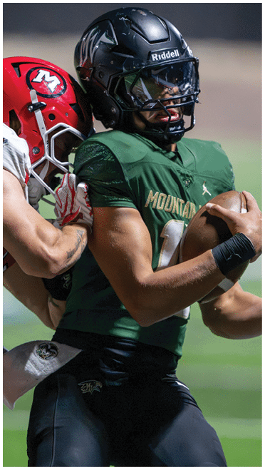 Two people in football uniforms are engaged in a close physical play on a field, with one wearing a red helmet and white jersey and the other in a dark helmet and green jersey. Description generated by AI