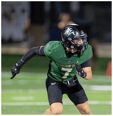 A person wearing a green football uniform and black helmet is in an athletic stance on a field. Description generated by AI