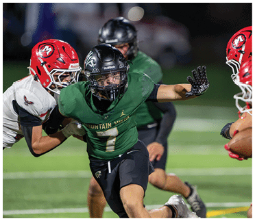 A person in a green football uniform is running while being closely pursued and grabbed by two people in white and red football uniforms on a field. Description generated by AI