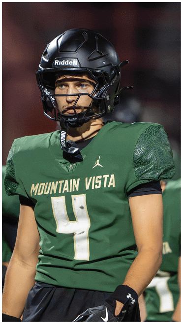 Two people wearing green football uniforms and black helmets are standing on a field, with one person in clear focus and the other blurred in the background. Description generated by AI