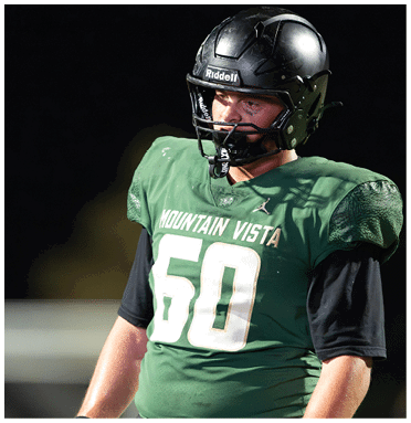 A person wearing a black football helmet and green jersey looks intently to the side against a dark background. Description generated by AI