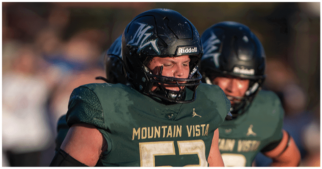 Two people wearing dark green football uniforms and black helmets with “Mountain Vista" on the jerseys are on a field during a game. Description generated by AI