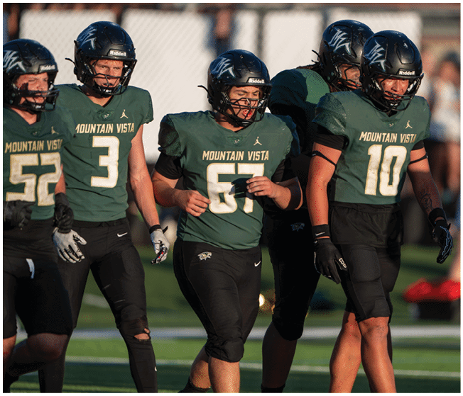 Five people wearing Mountain Vista football uniforms and helmets are walking together on a field during a game or practice. Description generated by AI