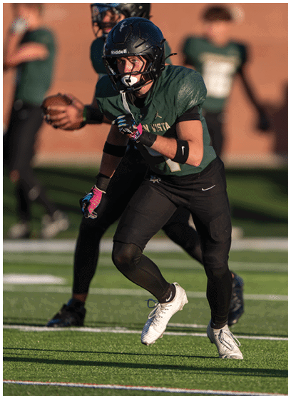 A person wearing a black football helmet and green jersey is in a ready stance on a football field with another person holding a football in the background. Description generated by AI