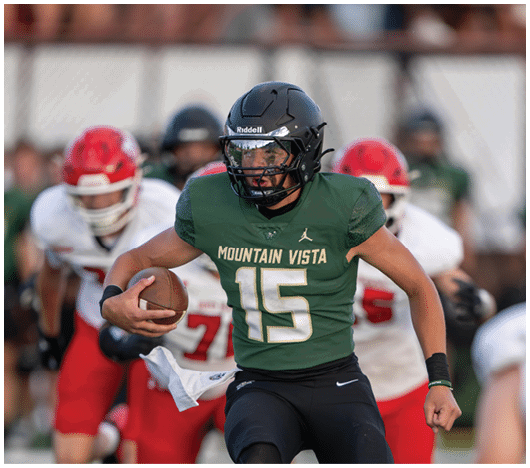 A person wearing a green Mountain Vista football jersey and black helmet is running with a football while other players in red and white uniforms are in the background. Description generated by AI