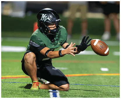 A person wearing a green and black football uniform and helmet is kneeling on one knee on a field while reaching out with both hands to catch a football. Description generated by AI