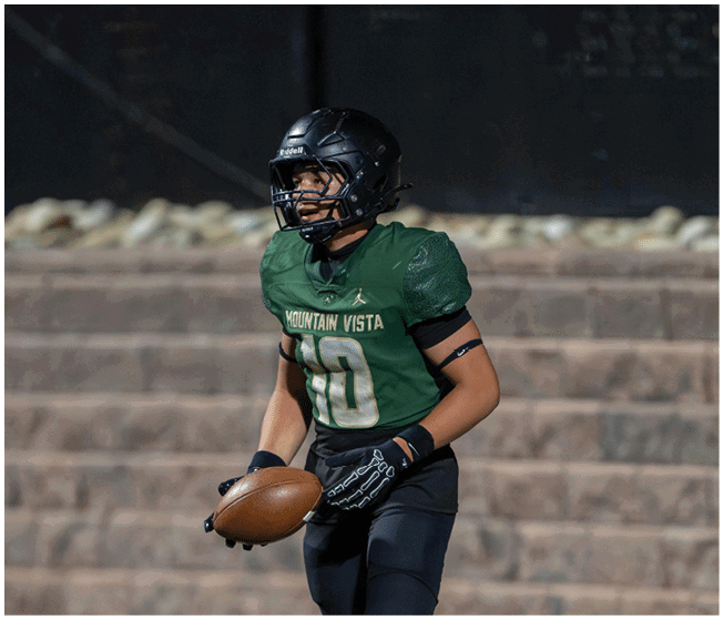 A person wearing a green Mountain Vista football jersey and black helmet is holding a football on a field with stone steps in the background. Description generated by AI