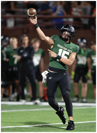 A person wearing a green Mountain Vista football jersey and black helmet is throwing a football on a field with teammates and coaches blurred in the background. Description generated by AI