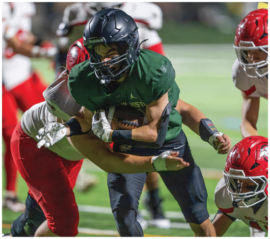 A person in a green football uniform and helmet is running with the ball while being tackled by a person in a white and red uniform on a football field. Description generated by AI