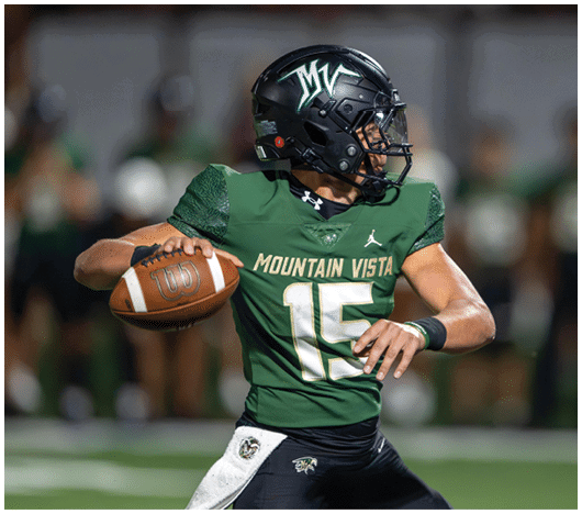 A person wearing a green Mountain Vista football uniform and black helmet is preparing to throw a football on a field. Description generated by AI
