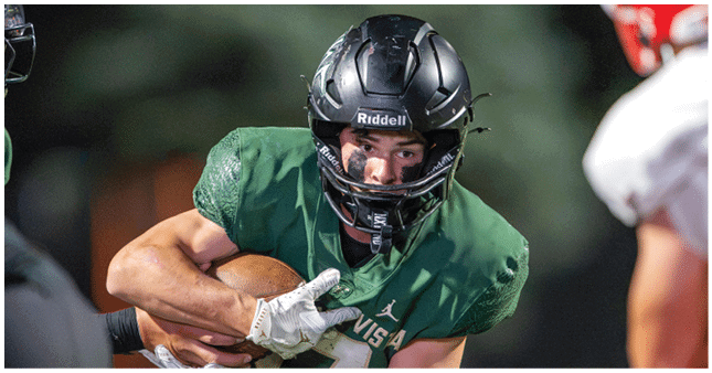 A person wearing a black helmet and green football uniform is holding a football tightly while running during a game. Description generated by AI