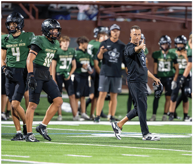 A group of people in green and black football uniforms stand on a field while a person in black athletic wear gestures with a thumbs-up. Description generated by AI