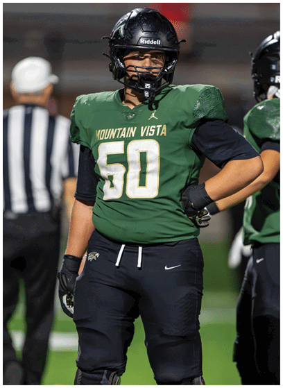 A person wearing a green Mountain Vista football jersey with the number 50 and black helmet stands on a field with hands on hips while another person and a referee are visible in the background. Description generated by AI