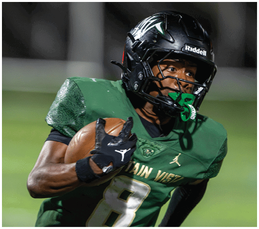 A person wearing a green football uniform and black helmet is holding a football and looking intently to the side. Description generated by AI