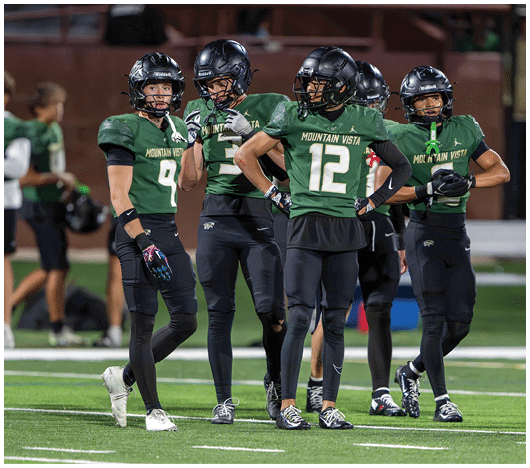 Two people in green football uniforms and helmets stand on a field while two others in similar attire are blurred in the background. Description generated by AI