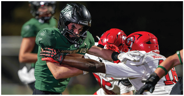 Two people in football gear are engaged in a physical struggle during a game, with one wearing a green uniform and the other in a white uniform with red helmets. Description generated by AI