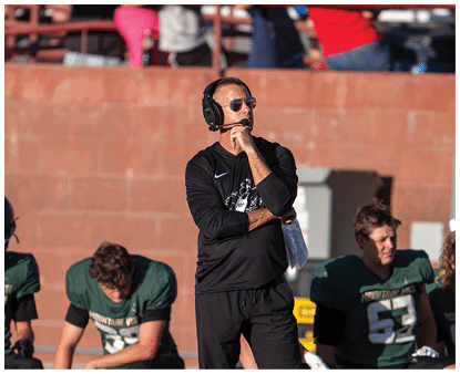 A person wearing a black long-sleeve shirt, sunglasses, and a headset stands with one hand on their chin and the other holding a clipboard, with people and a brick wall in the background. Description generated by AI