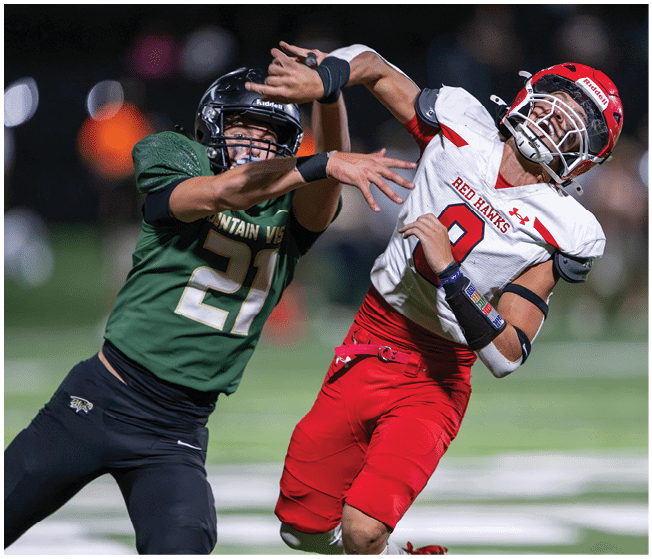 Two people in football uniforms are engaged in a dynamic play on a field, with one wearing a green jersey and the other in a white and red jersey. Description generated by AI