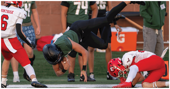 Three people wearing green football uniforms and black helmets stand on a field with blurred spectators in the background. Description generated by AI