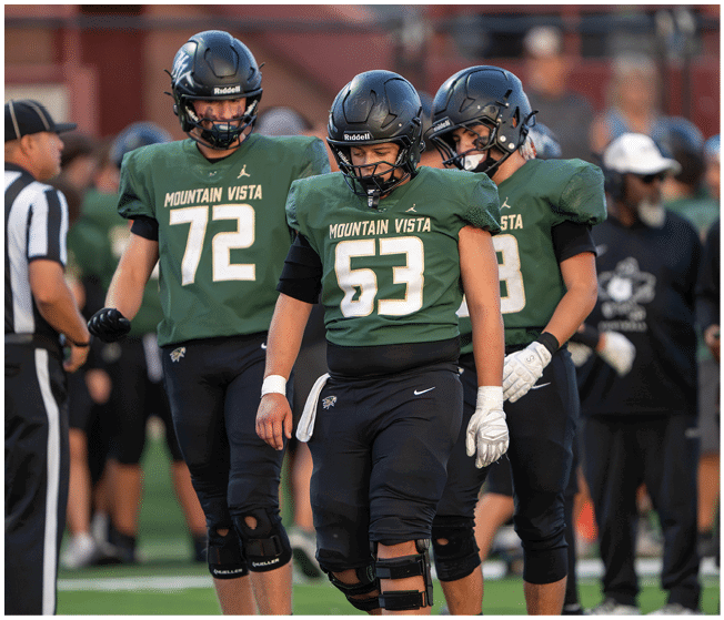 Three people in green Mountain Vista football uniforms and helmets stand on a field near a referee wearing a black and white striped shirt. Description generated by AI