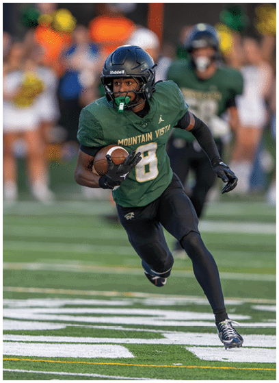 A person wearing a green football uniform with the words Mountain Vista is running on a field while holding a football. Description generated by AI