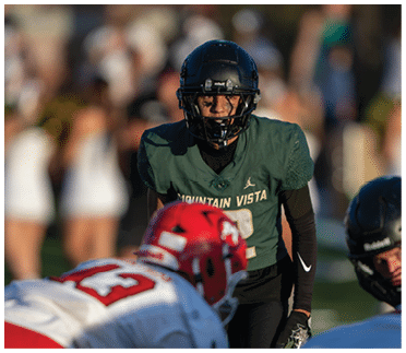 A person in a dark green football uniform with “Mountain Vista" on the jersey is focused and ready in a defensive stance facing another person in a white and red football uniform on a field. Description generated by AI