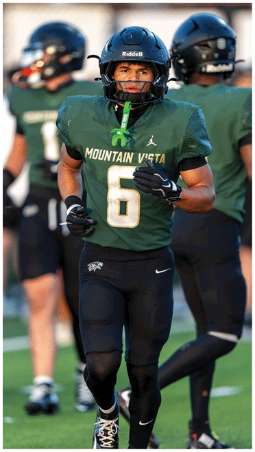 A person wearing a green Mountain Vista football uniform with the number six is running on a field with two other players in the background. Description generated by AI