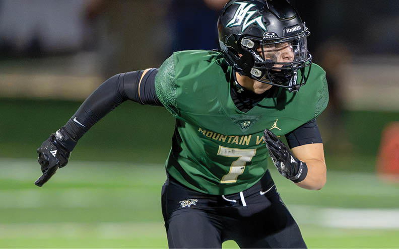 A person wearing a green football jersey and black helmet is in a ready stance on a football field. Description generated by AI