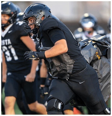 A person wearing a black football uniform and helmet is leaning forward, appearing focused and engaged in a game. Description generated by AI