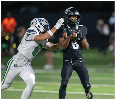 A person wearing a black football uniform and helmet is holding a football while another player’s hands reach toward them from behind. Description generated by AI