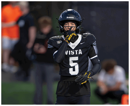 A person wearing a black football uniform with the number five and a helmet is holding a hand near their face while standing on a field. Description generated by AI