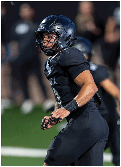A person wearing a black football uniform and helmet is running on a field during a game. Description generated by AI