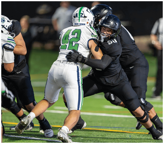 Two people in football uniforms are engaged in a physical block on a field during a game, with one wearing white and the other in black. Description generated by AI