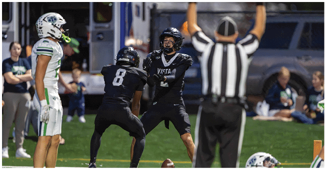 Two people in black football uniforms are preparing to catch or throw a ball while a referee raises both arms and a person in a white football uniform watches nearby. Description generated by AI