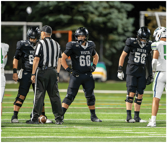 Three people in black football uniforms with the word Vista stand on a field with a referee and a person in a white uniform during a game. Description generated by AI