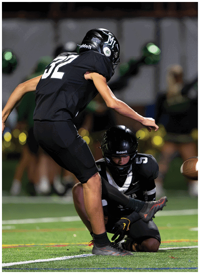Two people in black football uniforms are on a field with one person kneeling and holding a football while the other is preparing to kick it. Description generated by AI