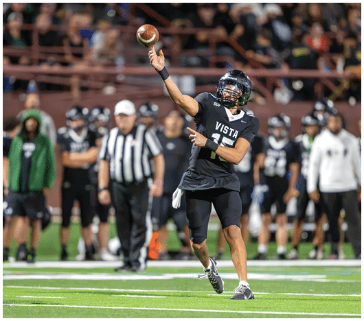 A person wearing a black football uniform with the word Vista is throwing a football on a field with other players and a referee in the background. Description generated by AI