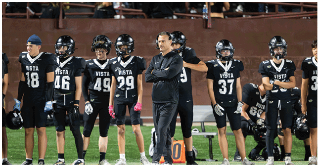 Adults in black football uniforms with “VISTA" on the jerseys stand and sit on the sidelines of a football field with one adult in black athletic wear standing with arms crossed. Description generated by AI