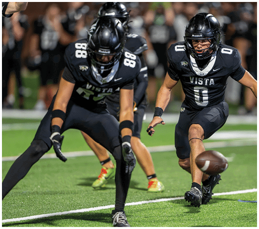 Two people in black football uniforms with “VISTA" on their jerseys are actively engaged in a play on a green field, with one person reaching toward a football on the ground. Description generated by AI