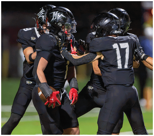 Four people in black football uniforms and helmets are gathered closely on a field, with one holding a football. Description generated by AI