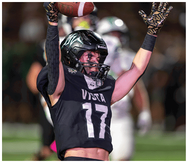 A person wearing a black football uniform with the number 17 and a helmet is raising their arms in celebration on a football field. Description generated by AI