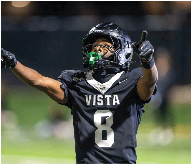 A person wearing a black football jersey with the word VISTA and the number 8 is raising their arms on a blurred outdoor field. Description generated by AI