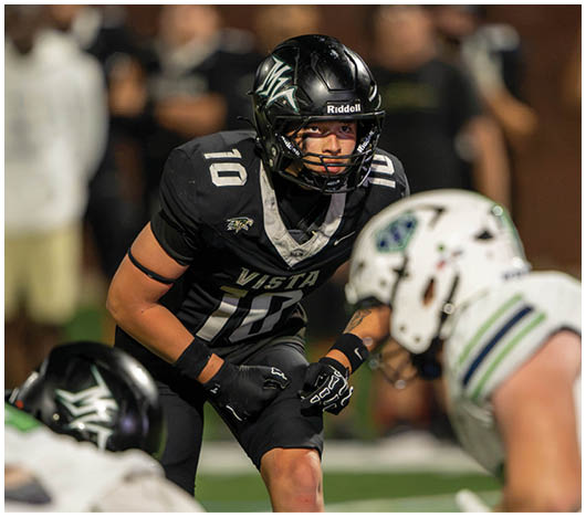 An athlete in a black football uniform with the number 10 is crouched and focused on the opposing team in white uniforms during a game on a green field. Description generated by AI