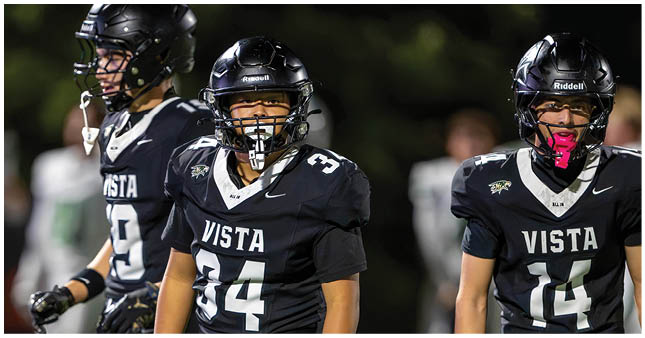 Three adults in black football uniforms with the word Vista on the front stand on a field during a game. Description generated by AI