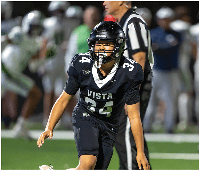 A person in a black football uniform with the number 34 and the word Vista is on a field, appearing focused and ready to move, while other players and a referee are blurred in the background. Description generated by AI