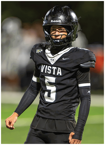 A person wearing a black football uniform with the word Vista and the number 5 on the jersey is standing on a field with a helmet and smiling. Description generated by AI