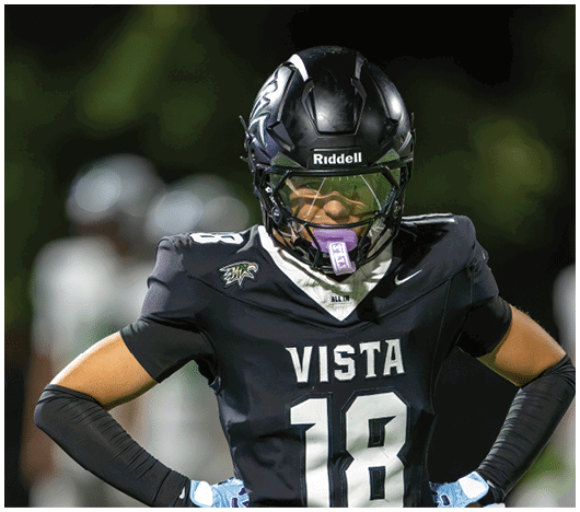 A person wearing a black football uniform with the number 18 and the word Vista stands with hands on hips on a field at night. Description generated by AI