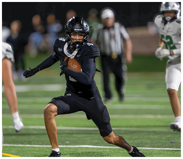 A person in black football gear is running on a field while holding a football, with other players and a referee visible in the background. Description generated by AI