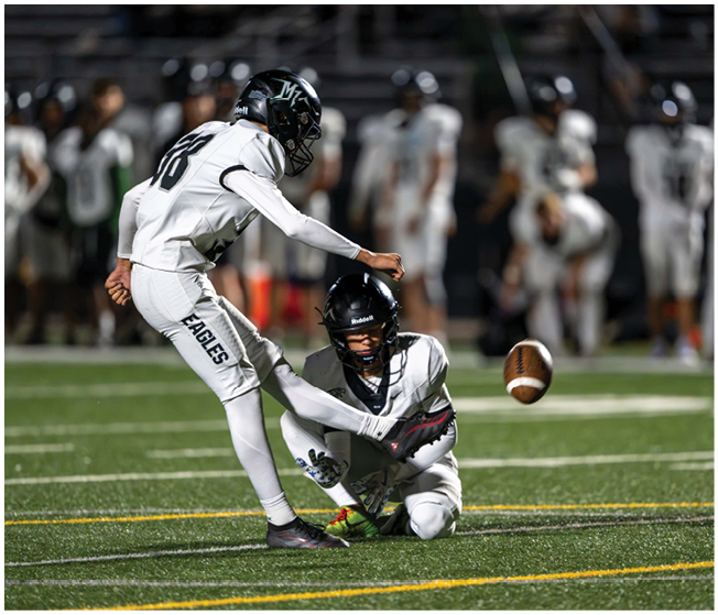 Two people in football uniforms are on a field with one holding the ball in place while the other kicks it during a game. Description generated by AI