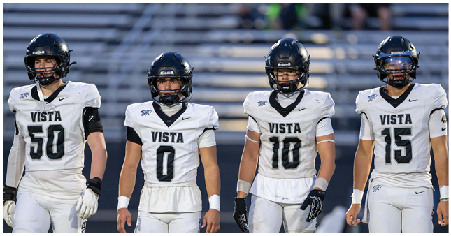 Four people wearing white football uniforms with black helmets and the word Vista on their jerseys stand side by side on a field with empty bleachers in the background. Description generated by AI